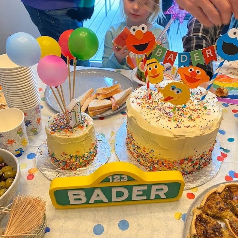 May include: A Sesame Street themed birthday cake with a white frosting and colorful sprinkles. The cake is decorated with Sesame Street characters and a banner that says "BADER". There is a smaller cake with a number one candle on top.