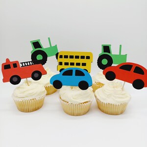 May include: Cupcakes topped with colorful vehicle cutouts, including a red fire truck, blue car, yellow school bus, and green tractors. The cupcakes have white frosting and are set against a white background.
