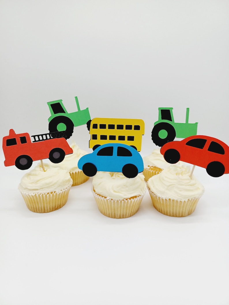 May include: Cupcakes topped with colorful vehicle cutouts, including a red fire truck, blue car, yellow school bus, and green tractors. The cupcakes have white frosting and are set against a white background.