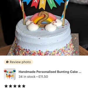 May include: A blue and white cake with colorful sprinkles around the bottom. The cake is decorated with a rainbow and a gold number 2 candle. A bunting with the name "ZAKARIYA" is on top of the cake. The bunting is made of colorful paper with Sesame Street characters Elmo and Cookie Monster on the ends.