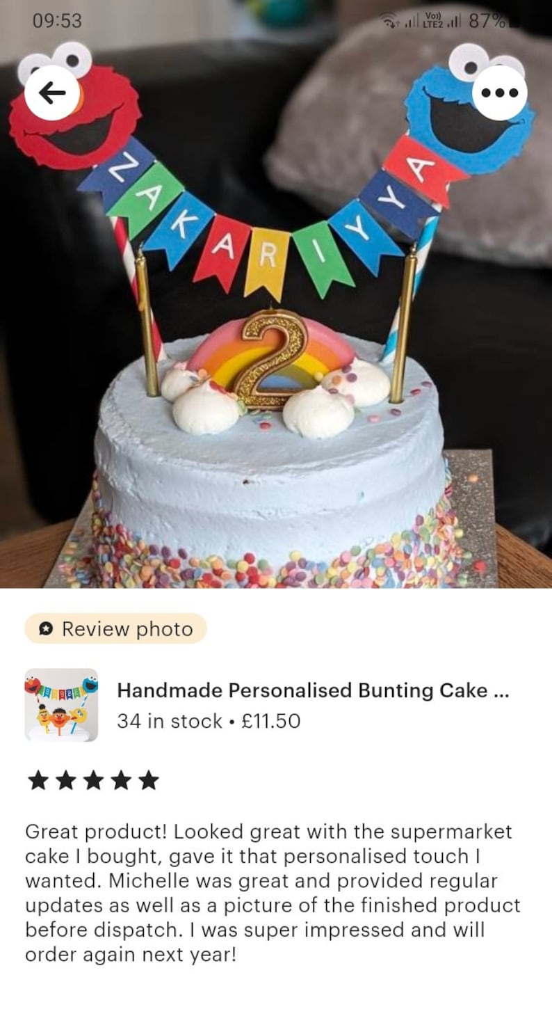 May include: A blue and white cake with colorful sprinkles around the bottom. The cake is decorated with a rainbow and a gold number 2 candle. A bunting with the name "ZAKARIYA" is on top of the cake. The bunting is made of colorful paper with Sesame Street characters Elmo and Cookie Monster on the ends.
