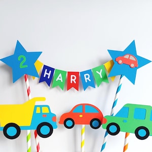 Bright primary coloured bunting cake topper with stripy straws.  2 stars either side of the bunting displaying the age in one star and a car in the other.  This topper comes complete with 3 vehicles.  This image shows a dumper truck & 2 cars.