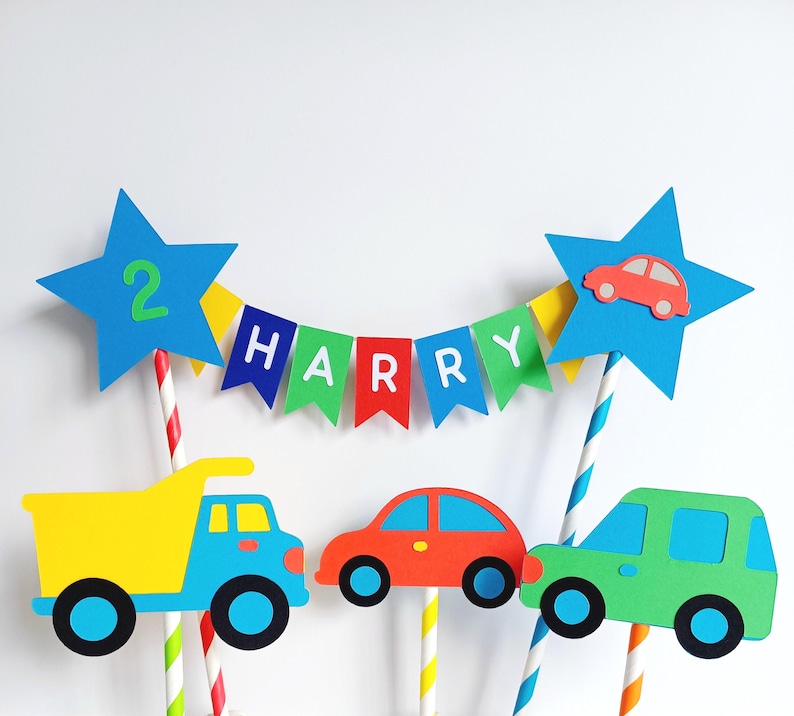 Bright primary coloured bunting cake topper with stripy straws.  2 stars either side of the bunting displaying the age in one star and a car in the other.  This topper comes complete with 3 vehicles.  This image shows a dumper truck & 2 cars.