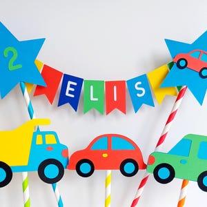 May include: A colorful birthday cake topper featuring a blue star with the number 2, a banner spelling "ELIS", and cutouts of a yellow dump truck, a red car, and a green SUV. The vehicles have black wheels and are attached to striped straws.