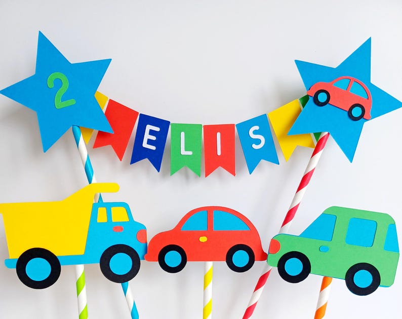 May include: A colorful birthday cake topper featuring a blue star with the number 2, a banner spelling "ELIS", and cutouts of a yellow dump truck, a red car, and a green SUV. The vehicles have black wheels and are attached to striped straws.