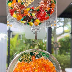 May include: A square glass suncatcher featuring a wreath of multicolored glass shards and the word Blessed in gold script, hanging above a circular glass suncatcher filled with orange glass pieces shaped like a pumpkin with green leaves.