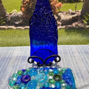 May include: A flattened cobalt blue glass bottle with a textured surface pattern displayed on a black metal stand, accompanied by a rectangular glass tray decorated with an assortment of blue and green glass beads and circular glass accents.