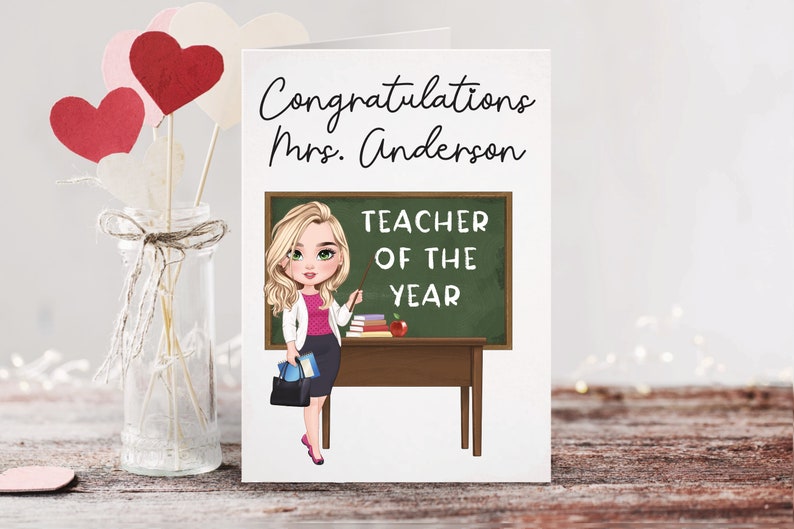 May include: A white card with the text "Congratulations Mrs. Anderson Teacher of the Year" and a cartoon illustration of a blonde woman standing in front of a chalkboard with the same text.