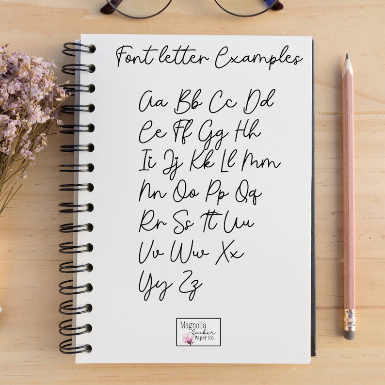 May include: A white notebook with a black spiral binding. The notebook has black handwritten text that says "Font letter Examples" and then lists the alphabet in cursive. The notebook also has a small sticker on the bottom right corner that says "Magnolia Ember Paper Co."