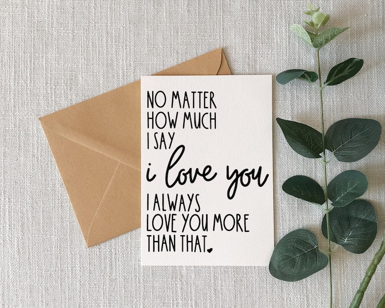 May include: A white greeting card with the text "NO MATTER HOW MUCH I SAY i love you I ALWAYS LOVE YOU MORE THAN THAT" in black script. A brown envelope is behind the card, and a sprig of greenery is to the right.