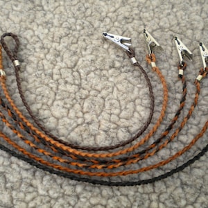 May include: A collection of six braided leather cord clips in various shades of brown and black. Each clip has a silver-colored metal clasp at one end and a loop at the other.