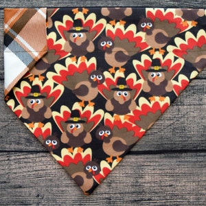 May include: A black bandana with a pattern of brown turkeys wearing pilgrim hats. The bandana has a red, orange, and white plaid trim.