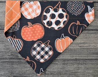 May include: Black and orange dog bandana with a pumpkin pattern. The bandana is made of fabric and has a triangular shape.