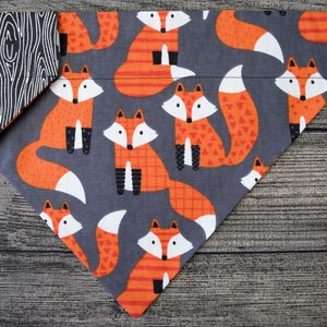 Op de afbeelding: Een grijze en oranje bandana met een vossenprint. De bandana is gemaakt van stof en heeft een zwart-wit houtnerfpatroon aan de achterkant.