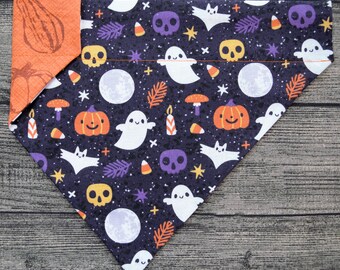May include: A black and white patterned fabric dog bandana with a Halloween theme. The bandana features ghosts, bats, pumpkins, candy corn, and stars. The bandana is trimmed with orange fabric.