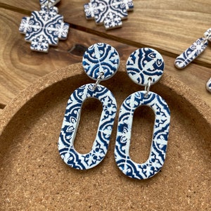 Könnte beinhalten: Weiß-blaue Ohrringe mit Muster in ovaler Form, die an silbernen Haken hängen. Weitere Ohrringe mit ähnlichem Design sind zu sehen, darunter kreuzförmige und runde Ohrstecker. Die Ohrringe werden auf einer Korkoberfläche präsentiert.