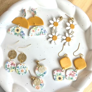 May include: A selection of handmade earrings on a white plate. The earrings showcase diverse designs, including daisy shapes, floral patterns, and geometric shapes in yellow, gold, and white. Some earrings have gold-coloured metal accents.