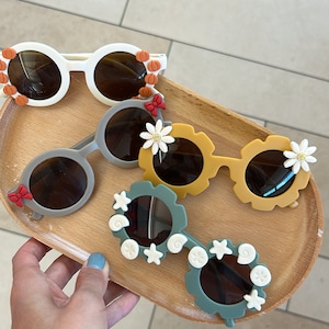 May include: Three pairs of colourful sunglasses with decorative details. The sunglasses are on a wooden tray. The sunglasses have round lenses and are made of plastic. The sunglasses are decorated with flowers, pumpkins, and seashells.