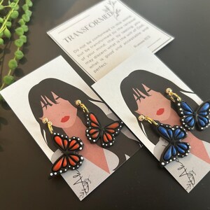 May include: Two pairs of butterfly earrings on display cards. One pair is black with red wings and the other is black with blue wings. The cards feature a stylized illustration of a woman with long hair.
