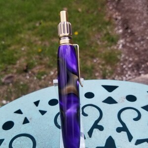 May include: A purple and gold pen with a swirl pattern on the barrel. The pen is on a clear acrylic stand.