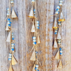 May include: A set of three gold-coloured wind chimes with blue and white beads hanging from a wooden surface. Each chime has multiple bells and is strung with beads.
