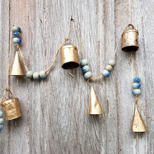 Op de afbeelding: Een reeks gouden bellen met blauwe en witte kralen die aan een houten oppervlak hangen. De bellen zijn van verschillende vormen en maten.