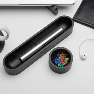 Pode incluir: Um porta-canetas preto com uma caneta prateada dentro, um pequeno recipiente preto com alfinetes coloridos e uma caneca de caf&eacute; branca com um descanso de copo preto.