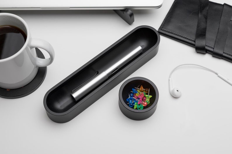 Pode incluir: Um porta-canetas preto com uma caneta prateada dentro, um pequeno recipiente preto com alfinetes coloridos e uma caneca de caf&eacute; branca com um descanso de copo preto.