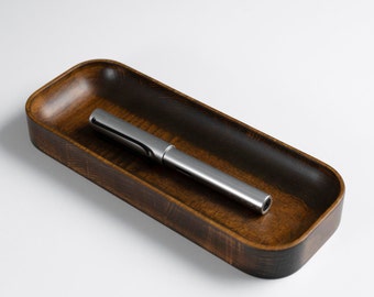 May include: A long, silver pen rests in a rectangular wooden tray. The tray has a dark brown stain and a smooth, polished finish.