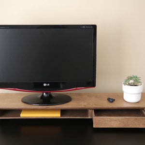 Puede incluir: Un soporte de monitor de madera con un cajón y un estante. El soporte es marrón y tiene un monitor negro en la parte superior. Hay una pequeña planta en maceta en el lado derecho del soporte.