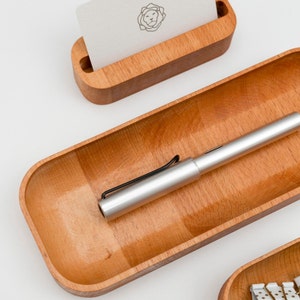 Op de afbeelding: Een houten dienblad met een zilveren pen erin. Het dienblad is gemaakt van licht hout en heeft een gladde afwerking. Er is een kleiner houten dienblad op de achtergrond met een witte kaart erin.