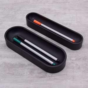 Pode incluir: Dois porta-canetas ovais pretos com acabamento liso e fosco. O porta-canetas superior tem uma caneta laranja dentro, e o porta-canetas inferior tem duas canetas, uma verde e uma preta.