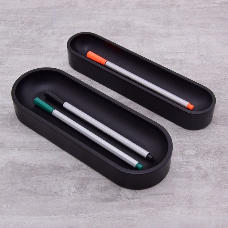 Pode incluir: Dois porta-canetas ovais pretos com acabamento liso e fosco. O porta-canetas superior tem uma caneta laranja dentro, e o porta-canetas inferior tem duas canetas, uma verde e uma preta.