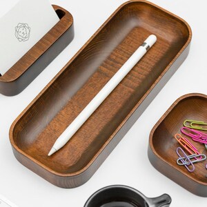 Op de afbeelding: Een houten dienblad met een witte stylus, een kleine houten kaarthouder, een kleine houten kom met kleurrijke paperclips en een zwarte koffiemok.
