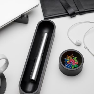 Pode incluir: Um porta-canetas preto com uma caneta prateada dentro, uma tigela preta com alfinetes coloridos e um cabo branco com fones de ouvido brancos.