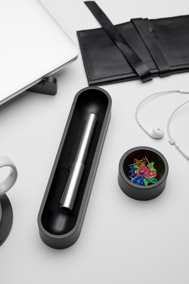 Pode incluir: Um porta-canetas preto com uma caneta prateada dentro, uma tigela preta com alfinetes coloridos e um cabo branco com fones de ouvido brancos.