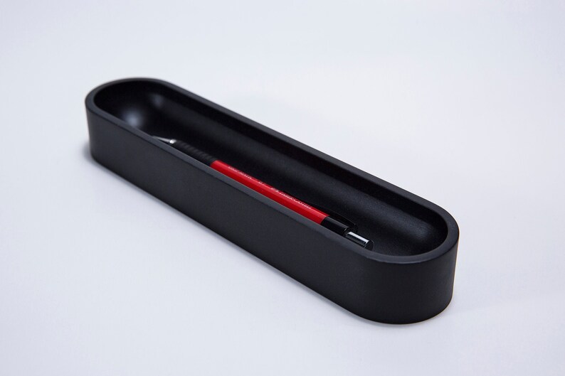 Pode incluir: Um porta-canetas preto de forma oval com uma caneta vermelha dentro.