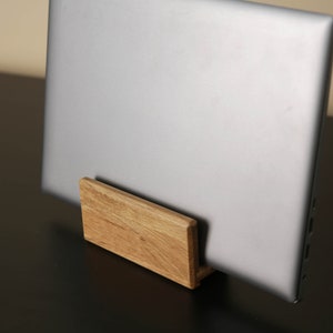 Pode incluir: Um laptop prateado está em um suporte de madeira. O suporte é feito de madeira clara e tem um design retangular simples. O laptop está fechado e inclinado, mostrando seu acabamento metálico elegante. A configuração sugere um espaço de trabalho minimalista.