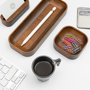 Op de afbeelding: Een wit bureau met een houten dienblad, een kleine houten kaarthouder, een zwarte koffiemok, een kleine vierkante houten kom met kleurrijke paperclips en een witte Apple Pencil. Het dienblad heeft een witte Apple Pencil erin. De kaarthouder heeft een witte kaart met een logo erin. De koffiemok is gevuld met zwarte koffie. De kom is gevuld met kleurrijke paperclips. Het bureau is wit en de achtergrond is wit.
