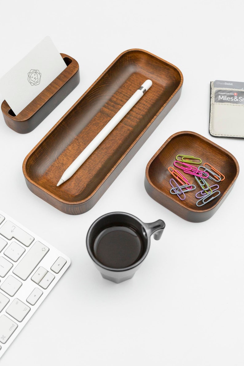 Op de afbeelding: Een wit bureau met een houten dienblad, een kleine houten kaarthouder, een zwarte koffiemok, een kleine vierkante houten kom met kleurrijke paperclips en een witte Apple Pencil. Het dienblad heeft een witte Apple Pencil erin. De kaarthouder heeft een witte kaart met een logo erin. De koffiemok is gevuld met zwarte koffie. De kom is gevuld met kleurrijke paperclips. Het bureau is wit en de achtergrond is wit.