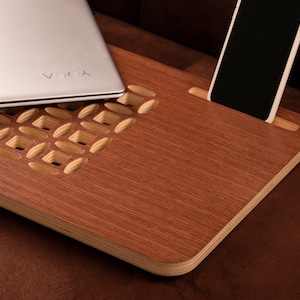 May include: A wooden laptop stand with a cutout design and a phone holder. The stand is brown with a light wood grain pattern. A laptop and a phone are placed on the stand.