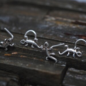 May include: Three silver nose rings shaped like lizards. The rings are on a dark brown wooden surface.