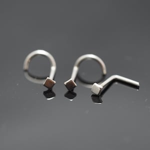 May include: Three silver nose rings with square-shaped ends. The rings are on a dark gray surface.