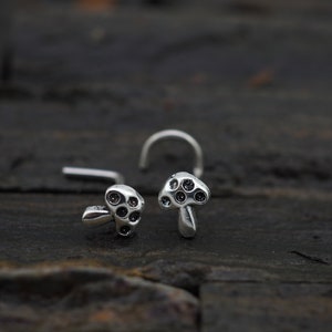 Puede incluir: Dos anillos de nariz de plata con diseños de hongos. Cada hongo tiene una parte superior redondeada con puntos negros y un tallo.