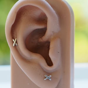 以下が含まれることがあります： マネキンの耳に、銀製のクロススタッドピアスが2つ。ピアスは小さく繊細で、シンプルなデザインです。