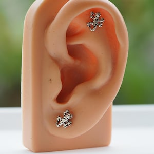 May include: Silver-toned lizard-shaped stud earrings with black accents are displayed on a mannequin ear. The earrings are small and detailed, showcasing a reptile design. The ear is a light peach color, set against a blurred background.