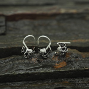 Puede incluir: Tres anillos de nariz de plata con diseños de calavera. Los anillos son de metal y tienen un pequeño bucle en la parte superior para sujetarse a un piercing en la nariz.