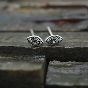 Puede incluir: Un par de pendientes de plata con forma de ojo y un diseño detallado. Los pendientes son pequeños y delicados, y serían un gran regalo para alguien que ama las joyas únicas.