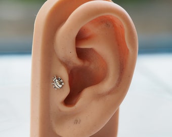 Puede incluir: Un pequeño pendiente de botón plateado con forma de mariquita se muestra en una oreja. El pendiente tiene detalles de patas y antenas. La oreja es de tono de piel claro y el fondo está borroso.