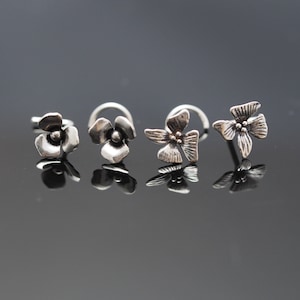 May include: Three silver nose rings with flower designs. The rings are all different shapes and sizes.
